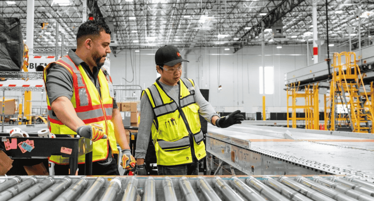 Two people in safety vests stand next to a conveyor. 