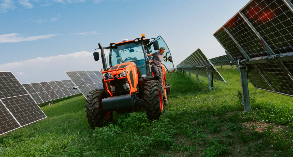 A person operates a tractor between solar panels in a green field. 