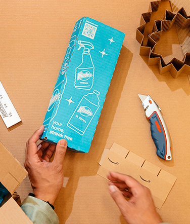 A hand is shown from above assembling a carboard box amidst tools on a table.