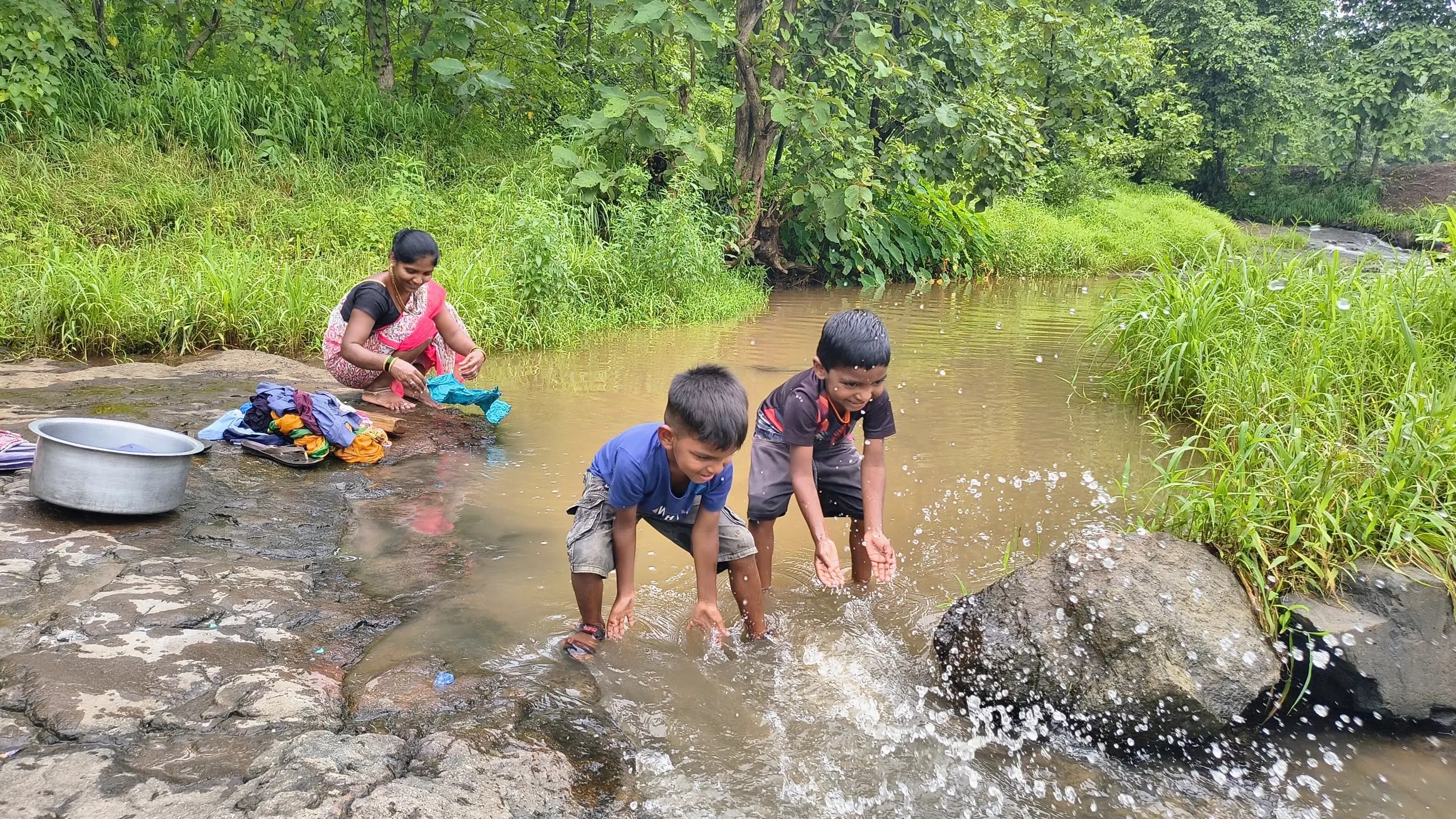 A woman washes clothes in a stream while two young children play in the water.