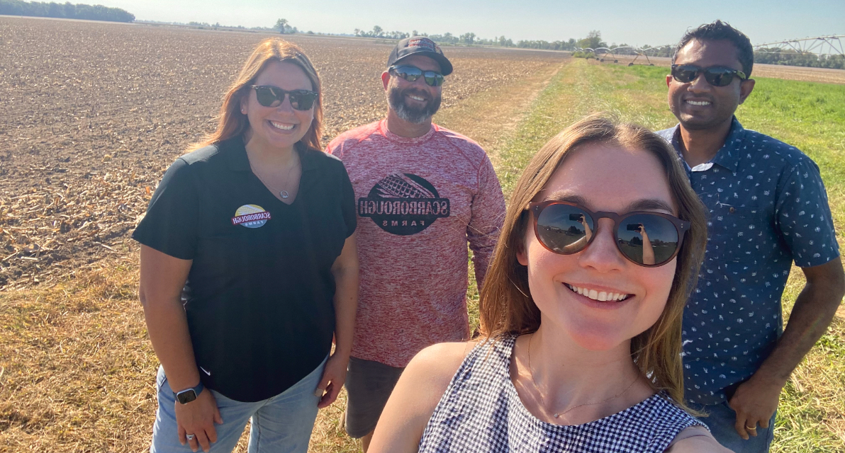 Four people gathered in a field smile for a photo. 